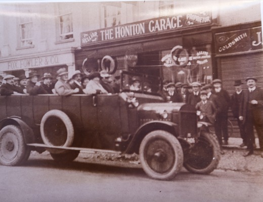 Sepia photograph of a charabanc outside The Honiton Garage with a crowd on the pavement