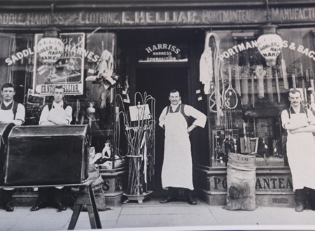 Sepia photograph of the old saddlery and harness shopfront with the proprietors standing outside in aprons