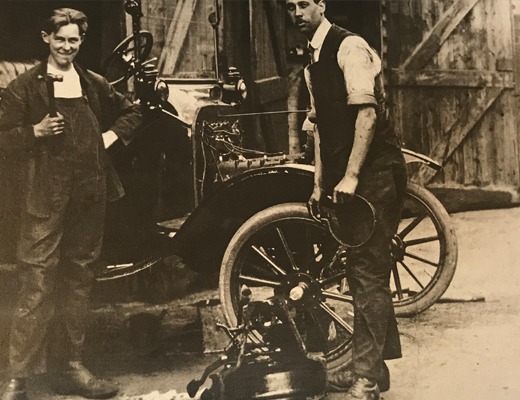 Sepia photograph of two men in work clothes beside an early motor car