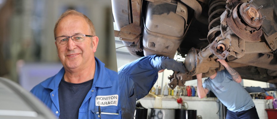Senior mechanic smiling while a colleague works under a raised car behind him